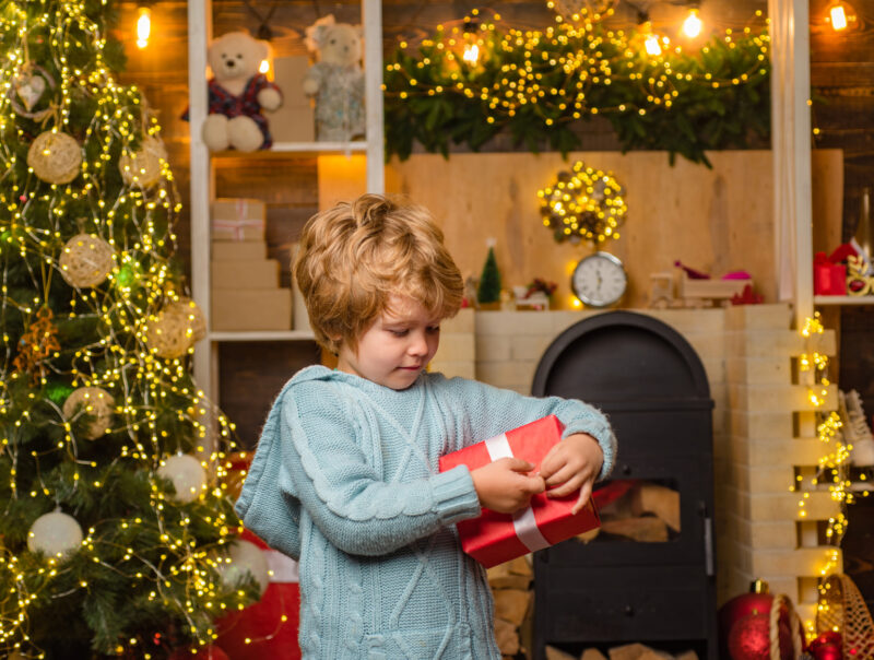 A child opening a Christmas present surrounded by a Christmas tree and fairy lights