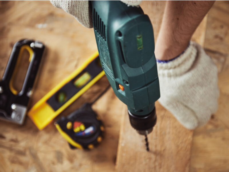 A man drilling a hole into a plank of wood with blurred DIY tools scattered in the background