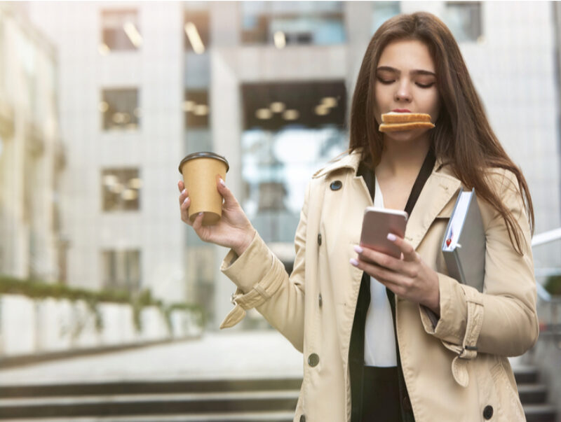 A young woman typing on a smartphone while holding a coffee and eating a sandwich