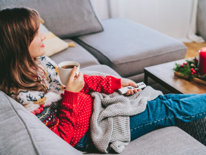 A woman in a Christmas jumper sits on the sofa with a cup of tea and a TV remote
