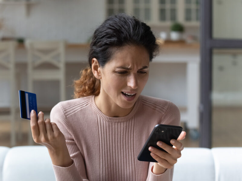 A stressed woman holding a credit card looks at her phone